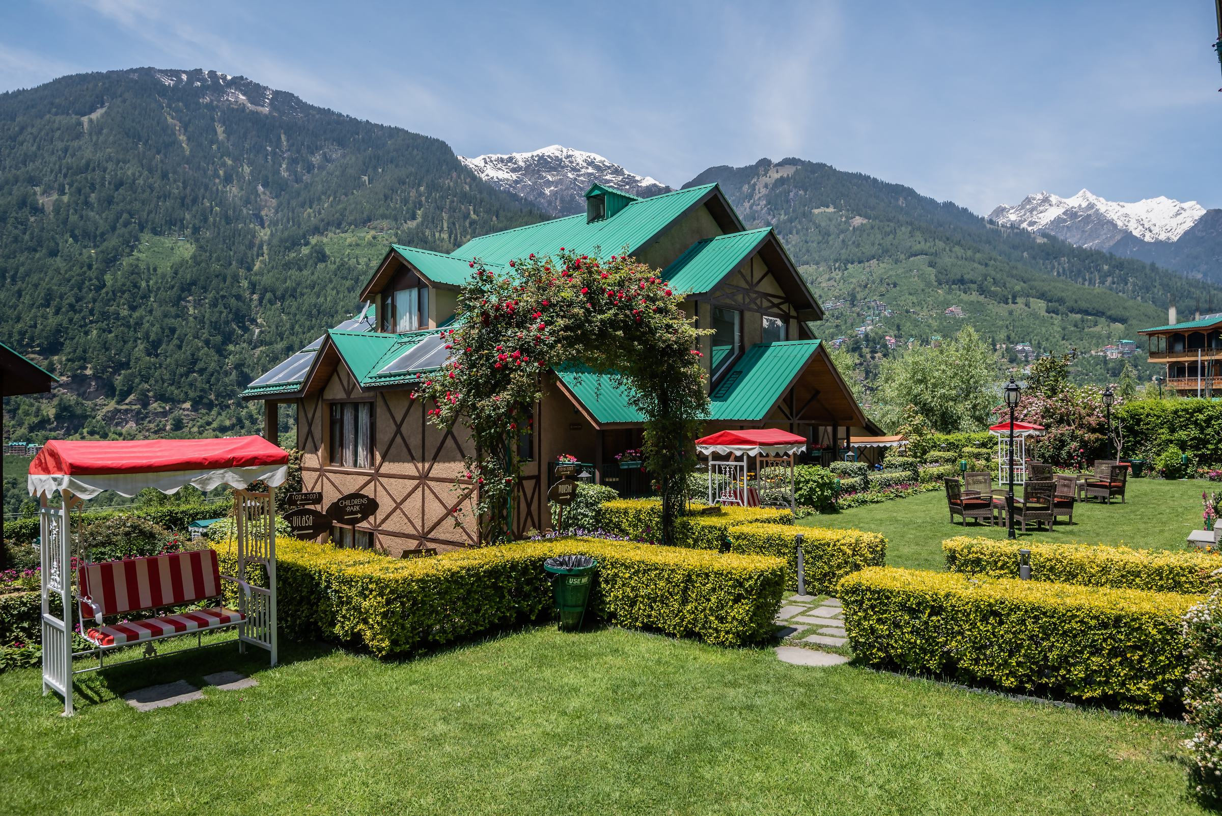 A green-roofed cottage behind a rose arch and clipped hedges, with snow peaks beyond